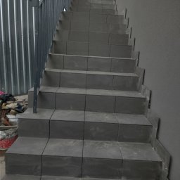 AK-BUD - Schody wyłożone szarymi płytkami ceramicznymi, widoczne narzędzia i wiadro po lewej stronie, metalowa barierka, ściana o szarej strukturze.