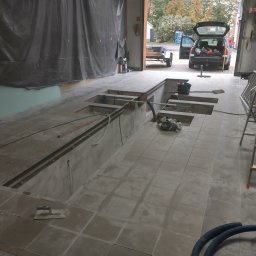 AK-BUD - Wnętrze pomieszczenia w trakcie prac remontowych, widoczna wykopana podłoga z fragmentami płytek, narzędzia murarskie, węże i drabina w tle otwarty garaż z zaparkowanym samochodem z przyczepą.