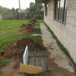 AK-BUD - Przygotowanie fundamentów pod taras betonowy przy ścianie budynku z białej cegły, widoczne wykopy z betonem, zbrojenie, szalunki z desek i styropianu, usypane pryzmy ziemi.
