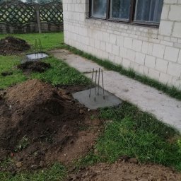 AK-BUD - Wylane betonowe stopy fundamentowe z wystającymi prętami zbrojeniowymi, widoczne pryzmy ziemi i fragment ściany budynku z białego bloczka.