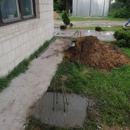 AK-BUD - Wylany betonowy fundament z wystającymi prętami zbrojeniowymi, obok ściana budynku z białej cegły i pryzma ziemi.