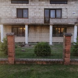 AK-BUD - Niedokończony dom z pustaków z balkonem i metalową balustradą, widoczny zza ogrodzenia z ceglanymi słupkami, z numerem domu '16' na ścianie.