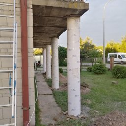 AK-BUD - Wzmocnione filary pod betonowym tarasem lub balkonem, zabezpieczone folią z nadrukiem, widok z boku na budynek z cegły i drabinę opartą o ścianę.