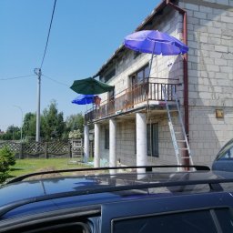 AK-BUD - Budynek z białej cegły z balkonami, na których ustawiono parasole przeciwsłoneczne; drabina oparta o balkon, widok zza samochodu.