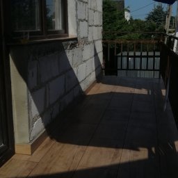 AK-BUD - Balkon z wyłożoną drewnopodobną terakotą, częściowo zacieniony, z widoczną ścianą z białego bloczka i oknem z szybą odbijającą zieleń drzew