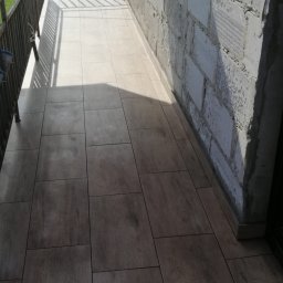 AK-BUD - Wyłożony płytkami balkon z surową ścianą z betonu komórkowego i metalową barierką, wpadające promienie słoneczne.