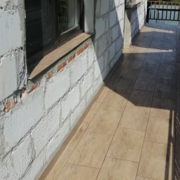 AK-BUD - Balkon w trakcie wykańczania: surowe ściany z betonu komórkowego, okno z brązową ramą, ułożona podłoga z płytek imitujących drewno, widoczny fragment balustrady i cień osoby fotografującej.