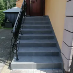 Zewnętrzne schody wejściowe wyłożone szarymi płytkami z czarną, kutą balustradą, prowadzące do brązowych drzwi wejściowych, obok ściana w kolorze piaskowym i kamienna kolumna.