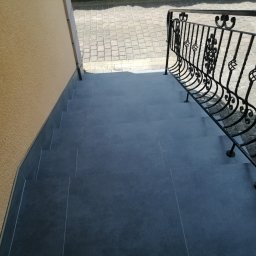 AK-BUD - Zewnętrzne schody wyłożone szarymi płytkami, z ozdobną, kutą, czarną balustradą, prowadzące do wejścia z kostki brukowej.