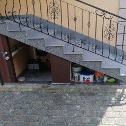 AK-BUD - Zewnętrzne schody z szarego granitu i jasnoszarych płytek, z ozdobną, kutą balustradą w kolorze czarnym. Pod schodami widoczne wiadro z farbą Bau, inne materiały budowlane i wejście...