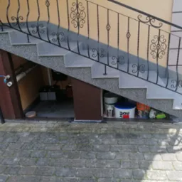 Zewnętrzne schody z szarego granitu i jasnoszarych płytek, z ozdobną, kutą balustradą w kolorze czarnym. Pod schodami widoczne wiadro z farbą Bau, inne materiały budowlane i wejście...