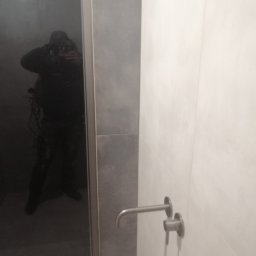 AK-BUD - Nowoczesna bateria umywalkowa zamontowana na jasnej, kafelkowanej ścianie, z widocznym odbiciem osoby robiącej zdjęcie w tafli szkła po lewej stronie.