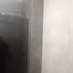 AK-BUD - Szklana kabina prysznicowa z deszczownicą, obudowana szarymi płytkami, przylegająca do białej ściany.