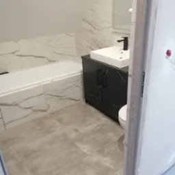 Ukończona łazienka na poddaszu z wanną obłożoną marmurowymi płytkami, umywalką nablatową na czarnej szafce i szarą podłogą z płytek, widok zza uchylonych drzwi.