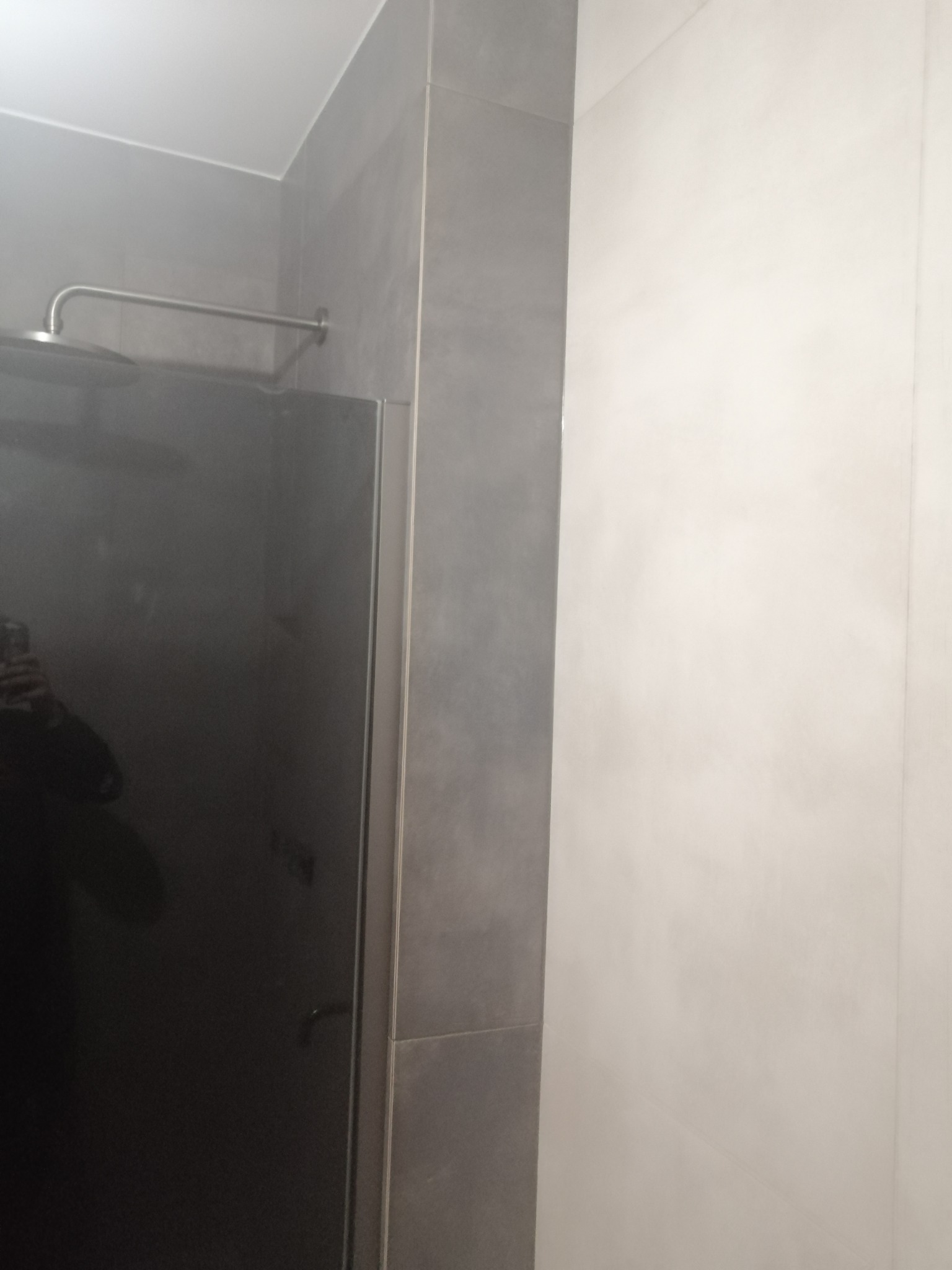 Szklana kabina prysznicowa z deszczownicą, obudowana szarymi płytkami, przylegająca do białej ściany.