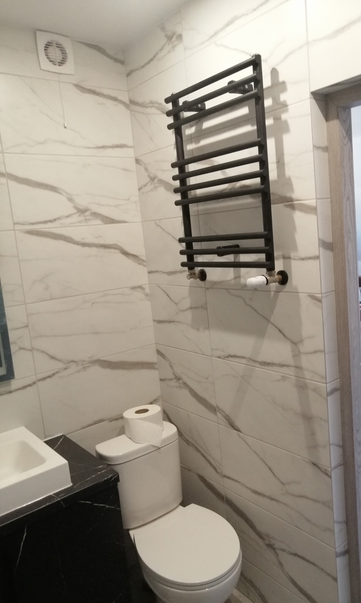 Nowoczesna łazienka z marmurowymi płytkami, czarnym grzejnikiem drabinkowym i toaletą z papierem toaletowym na górze.