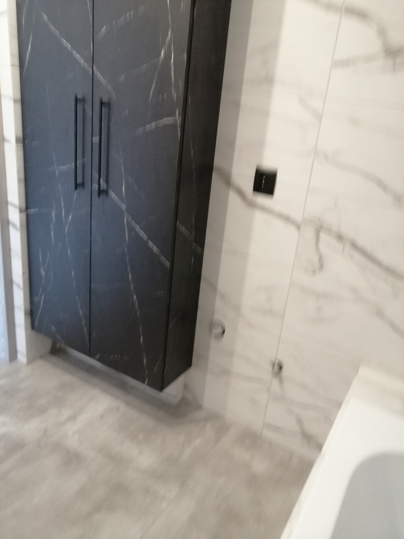 Szafka łazienkowa z czarnym frontem imitującym marmur, zamontowana na tle białych płytek marmurowych, widoczna część wanny.