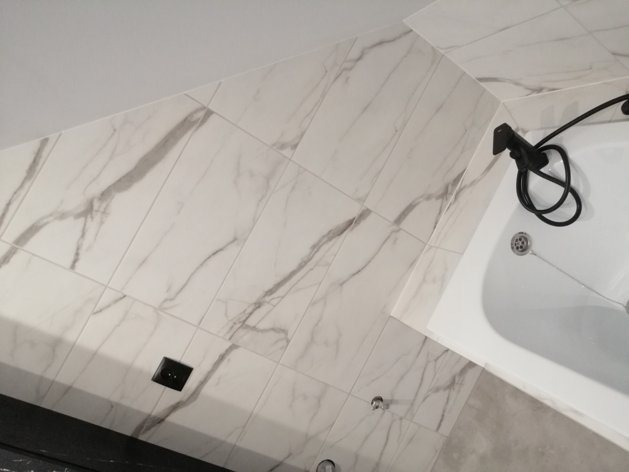Wykończona łazienka z białymi płytkami imitującymi marmur, czarną armaturą i odpływem liniowym w rogu pod skosem.