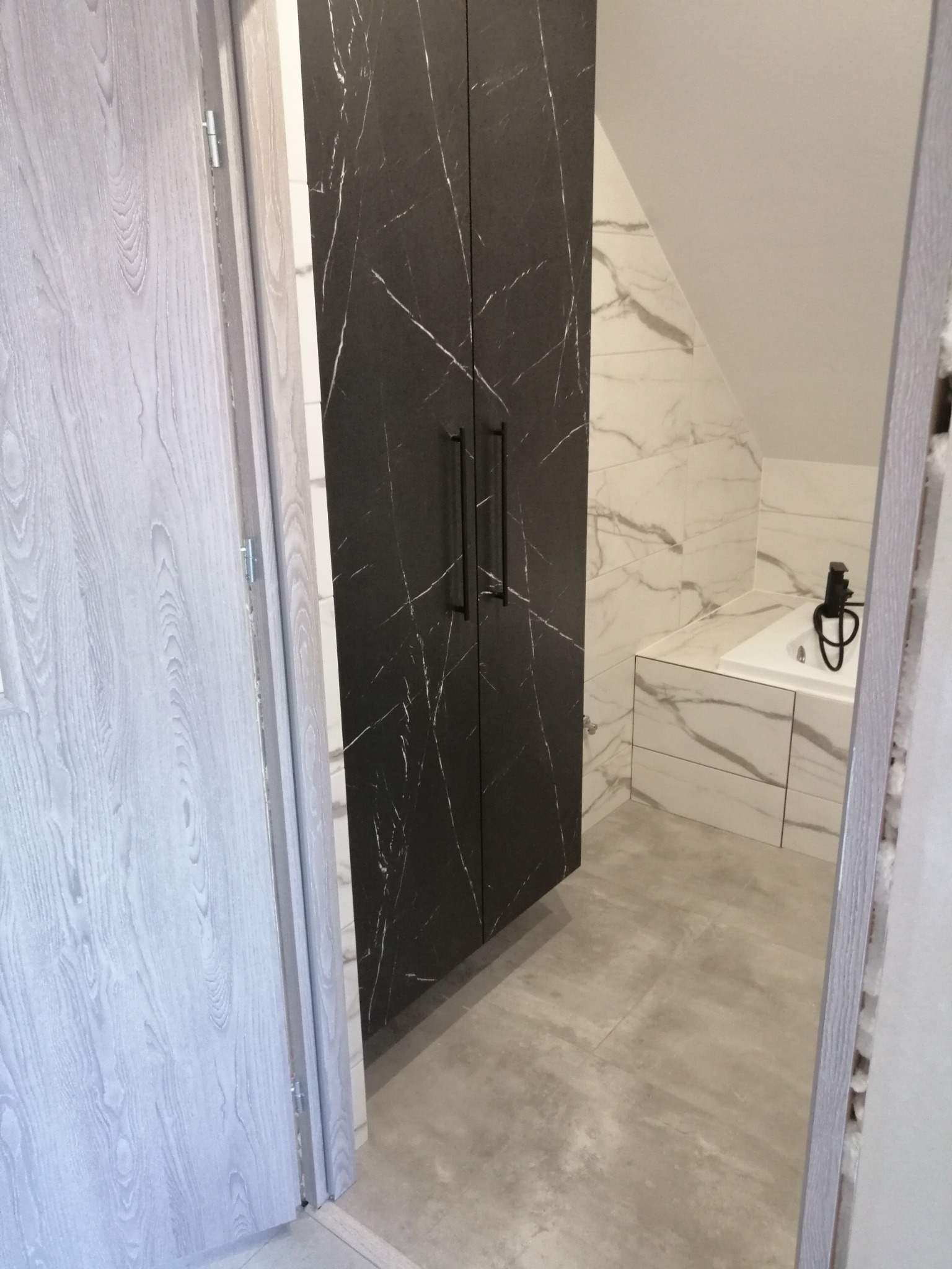 Wnętrze łazienki na poddaszu z zabudową szafy z czarnymi frontami imitującymi marmur, białe płytki z szarymi żyłami na ścianach oraz umywalka z czarną armaturą.