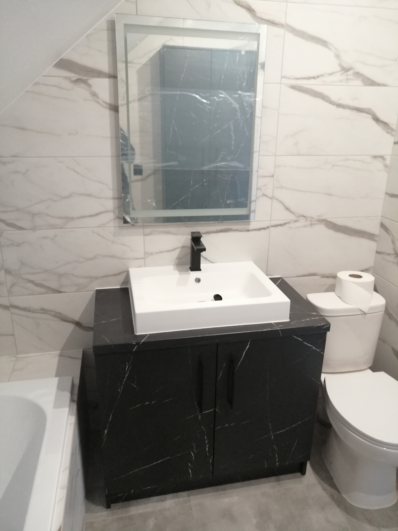 Nowoczesna łazienka z białą umywalką nablatową na czarnej szafce z marmurkowym wzorem, czarną baterią, podświetlanym lustrem i toaletą z papierem toaletowym na tle marmurowych płytek.