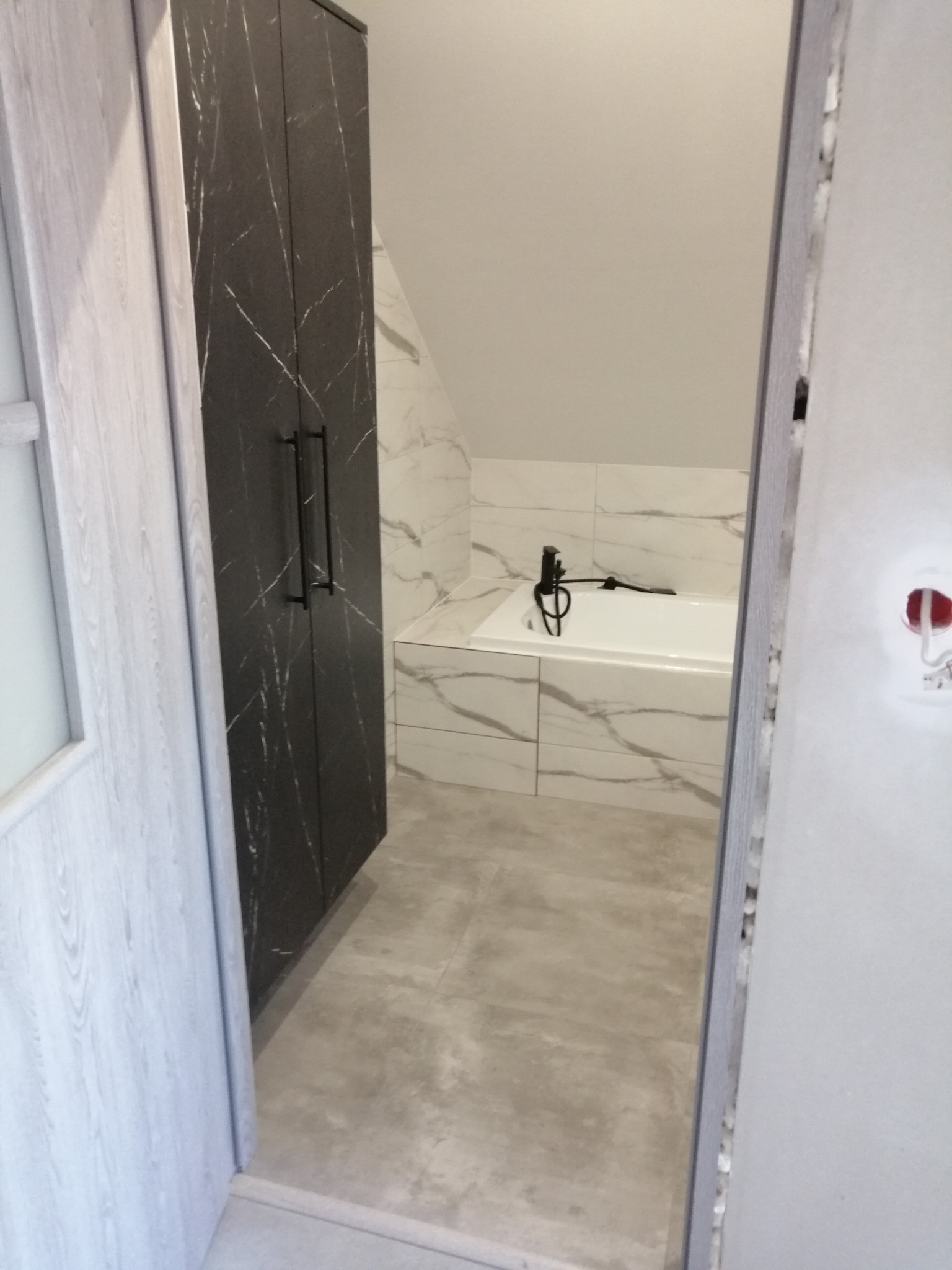 Nowoczesna łazienka w trakcie remontu: widoczna wanna z czarną armaturą, szafka w kolorze ciemnego marmuru oraz częściowo widoczne drzwi z ościeżnicą, z widocznymi śladami niedokończonych prac...