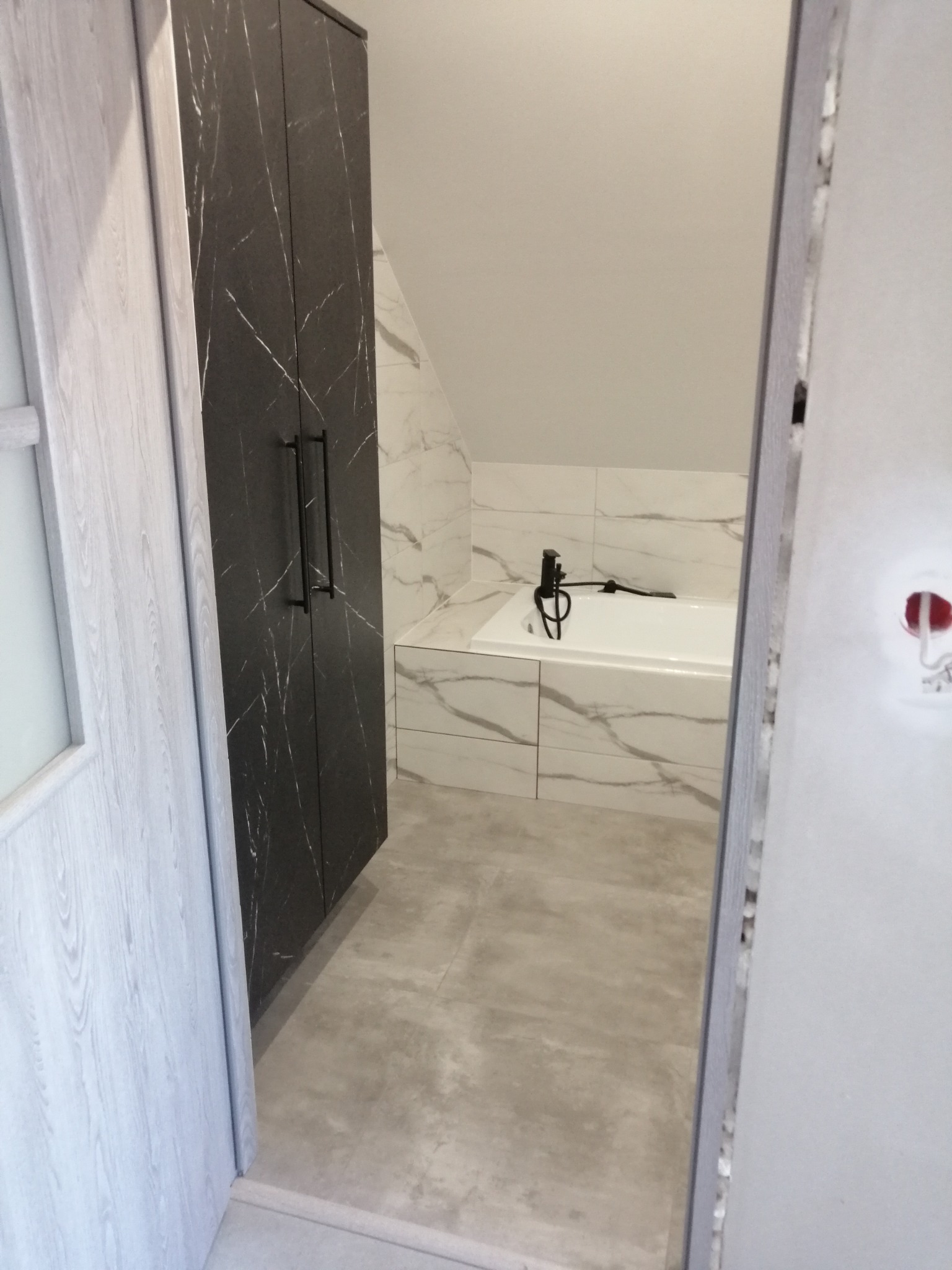 Wnętrze łazienki z wanną obłożoną marmurowymi płytkami, czarną armaturą i szafką z frontami imitującymi czarny marmur, widoczne przez futrynę drzwi.