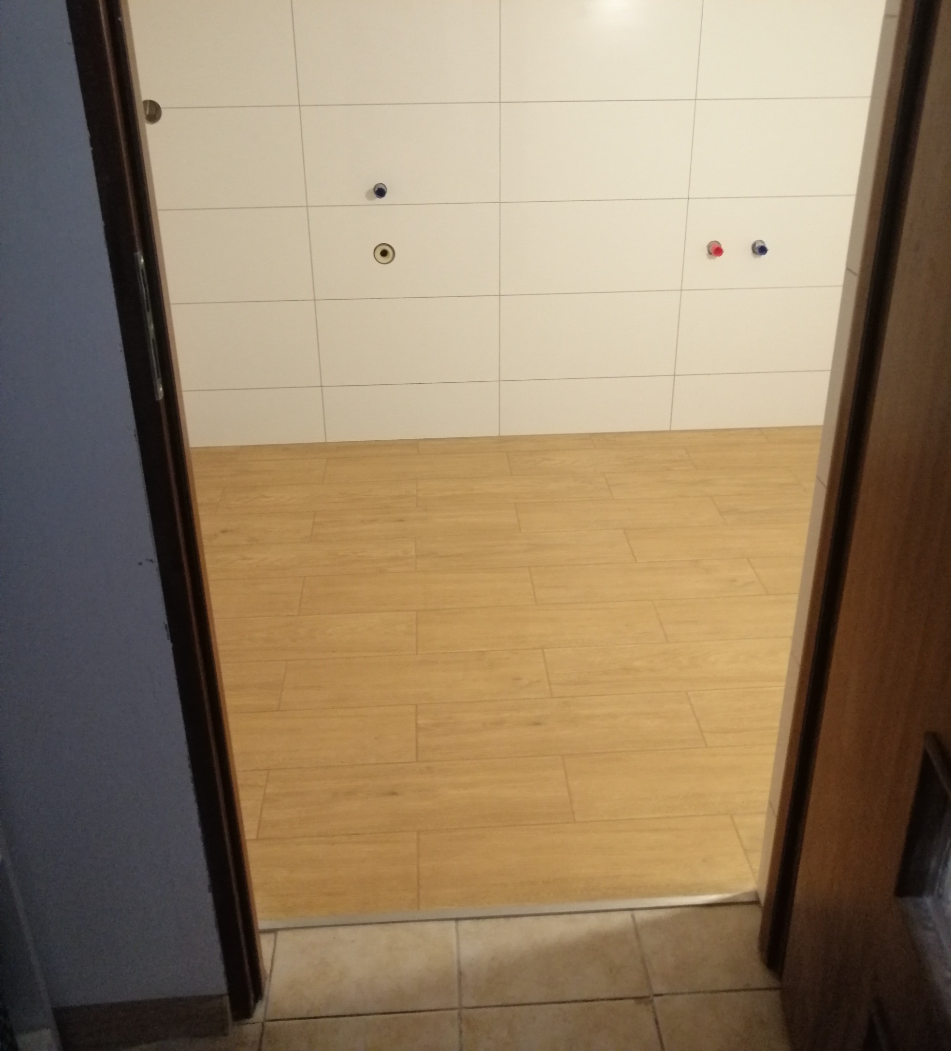 Wnętrze łazienki w trakcie remontu, widok zza uchylonych drzwi na świeżo położone płytki imitujące drewno na podłodze i białe płytki na ścianach z widocznymi przyłączami wodnymi.