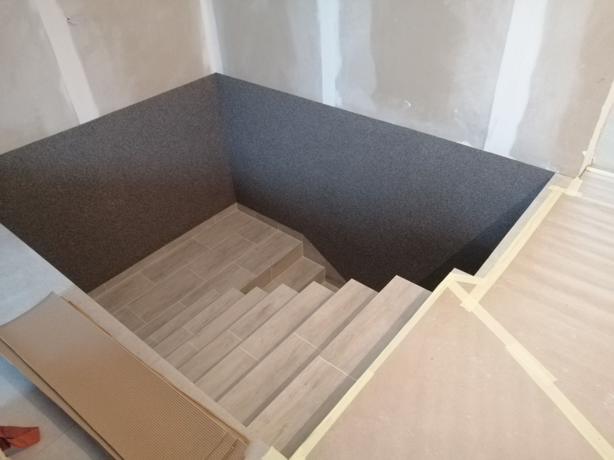 Schody wyłożone jasnymi płytkami w trakcie prac wykończeniowych, ściany w surowym stanie, zabezpieczone taśmą malarską.