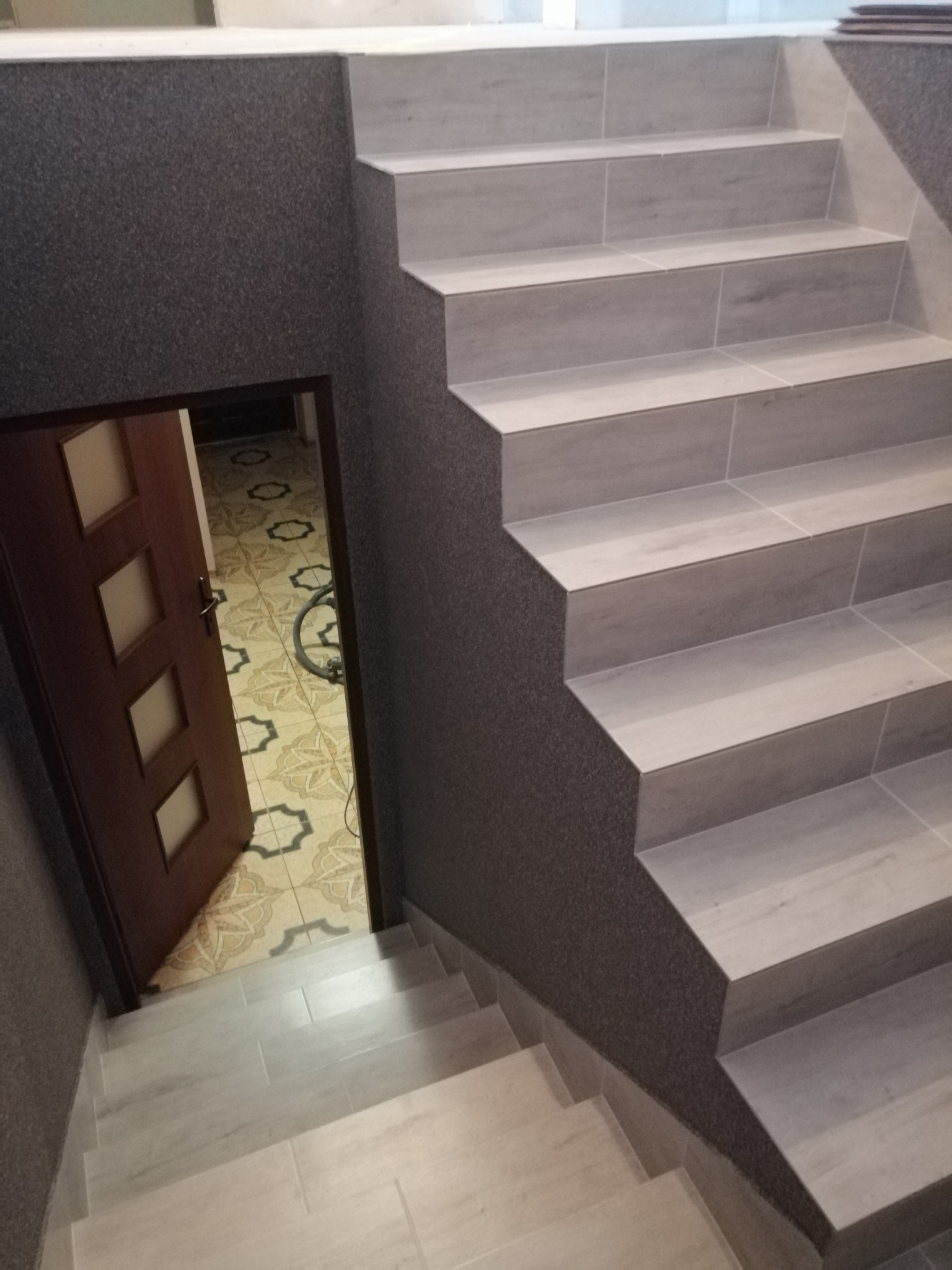 Wyłożone płytkami schody z szarymi ścianami prowadzące do otwartych drzwi z widokiem na wzorzystą podłogę.