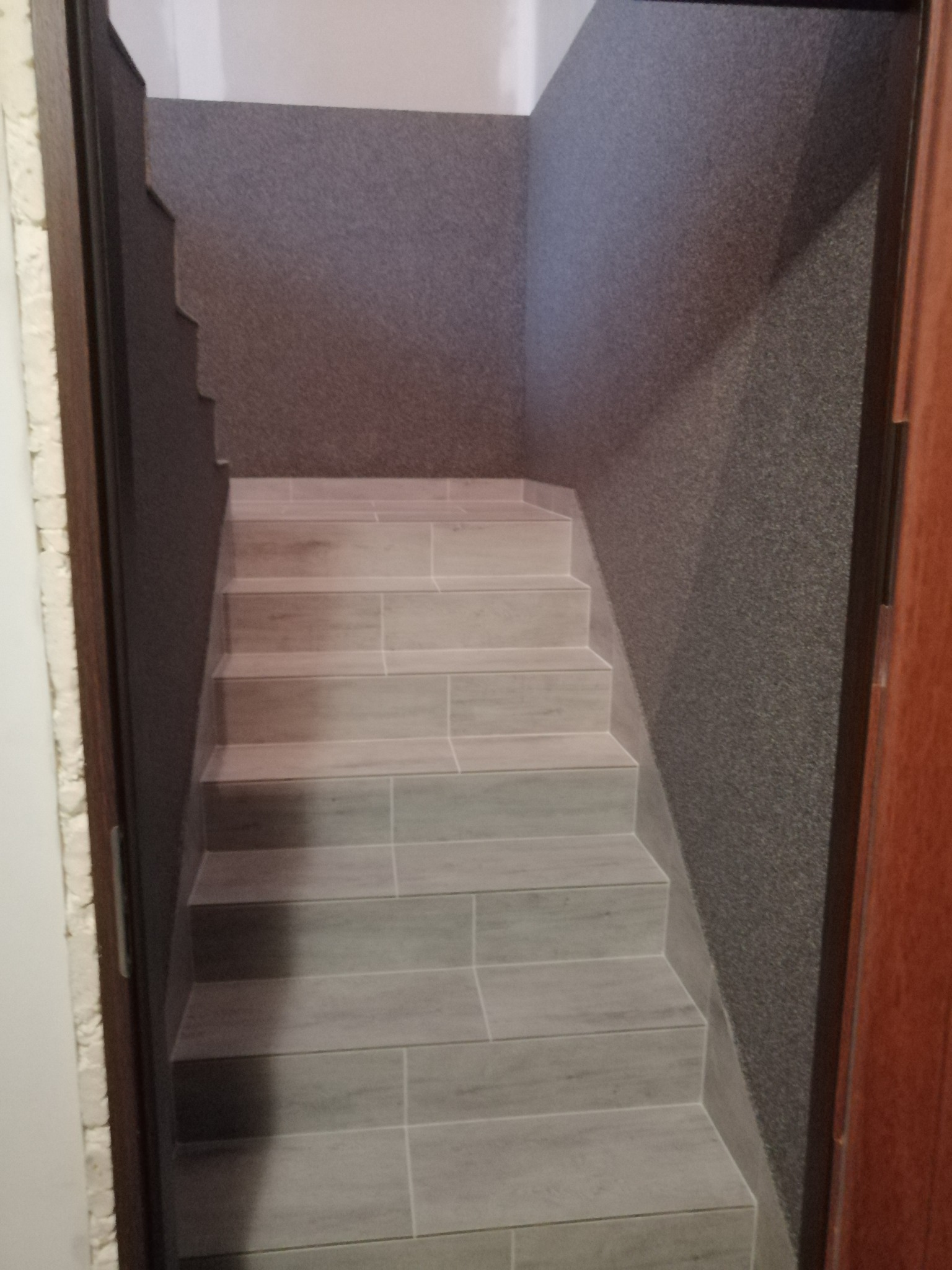 Wykończone schody wewnętrzne z szarymi płytkami imitującymi drewno, prowadzące w dół, z ciemną, teksturowaną ścianą po prawej stronie i widoczną ościeżnicą po obu stronach.