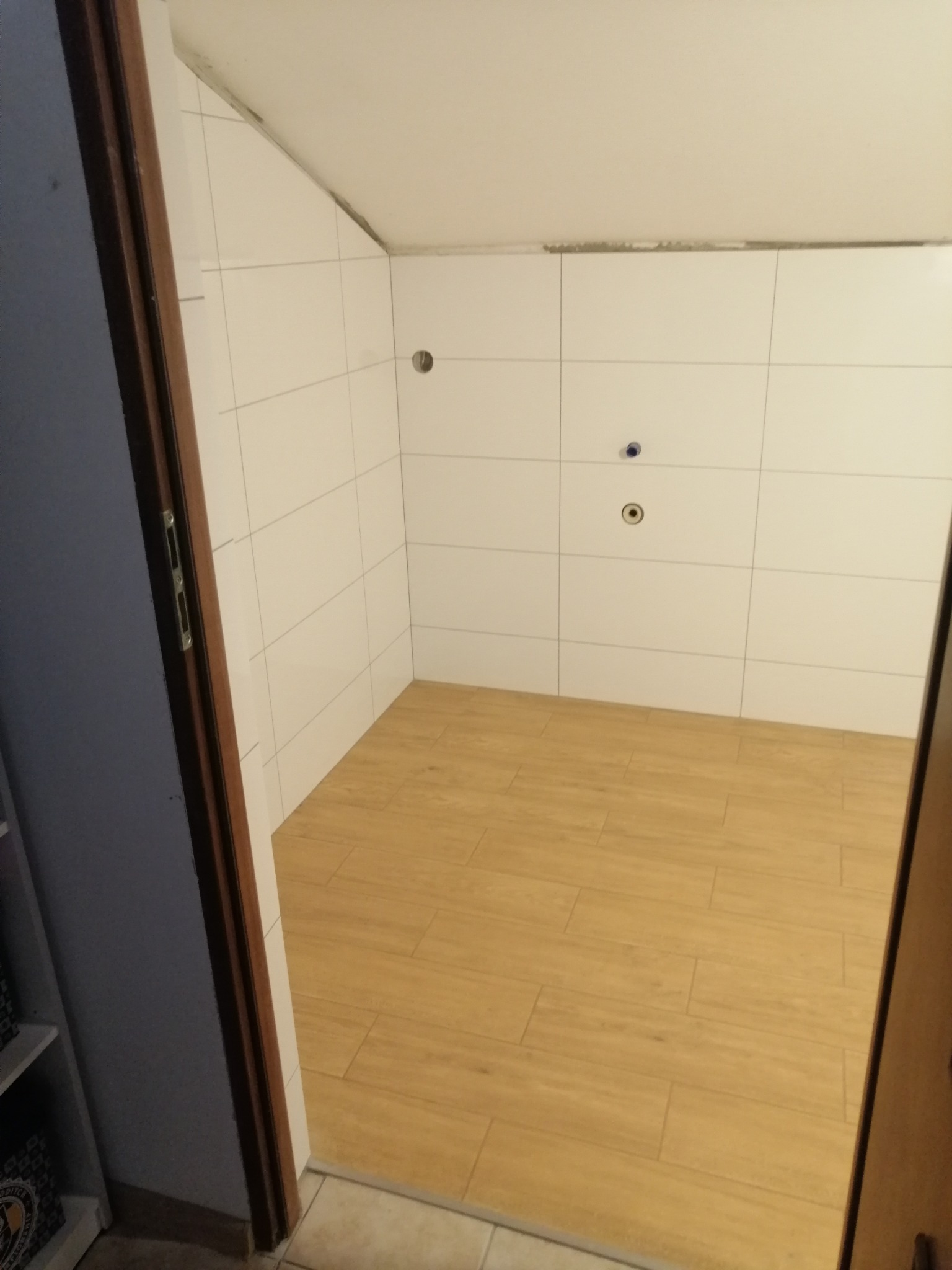 Pusta łazienka w trakcie remontu, wyłożona białymi płytkami ściennymi i drewnopodobnymi płytkami podłogowymi, widoczne otwory na instalacje.