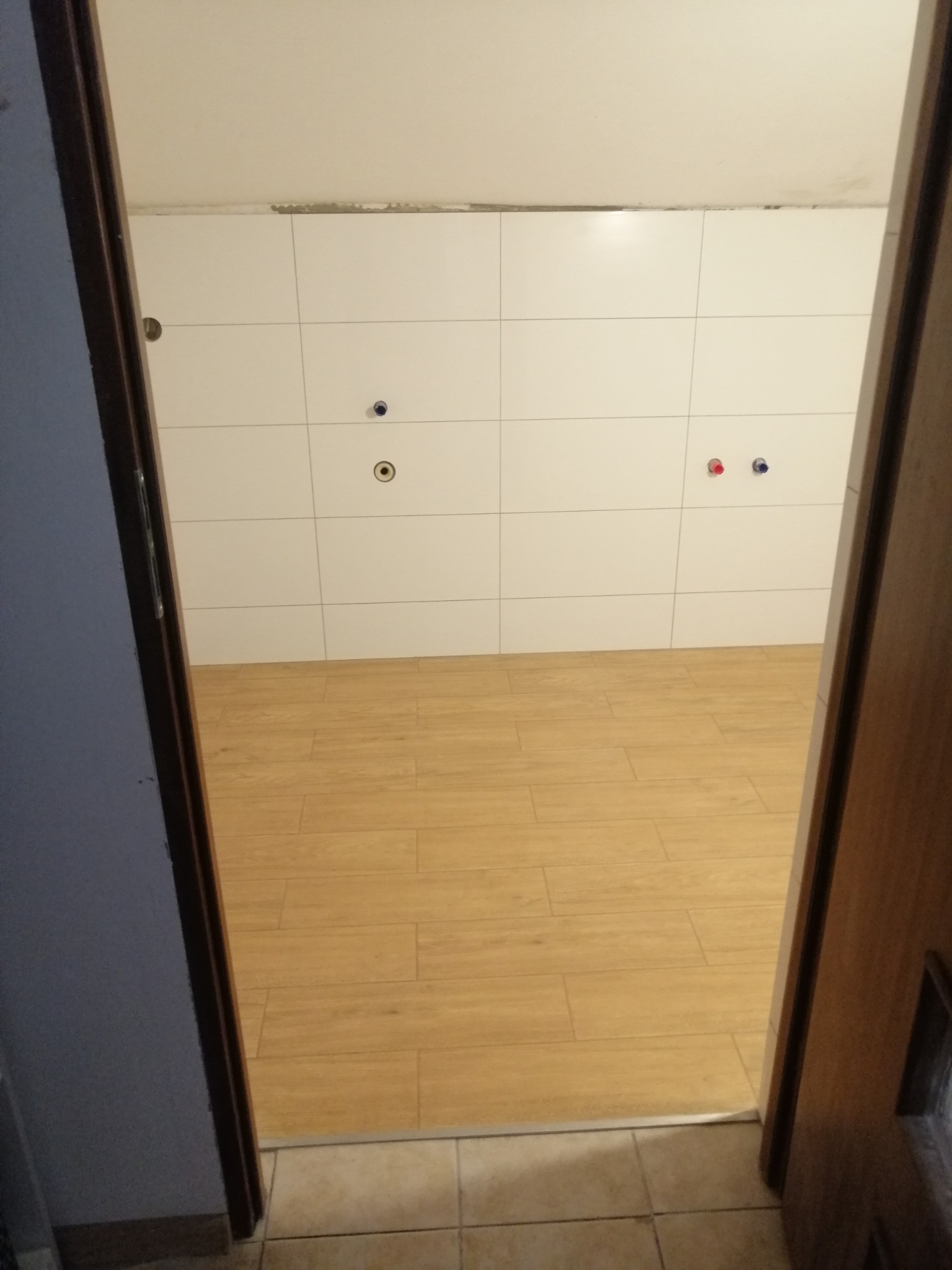 Wykończona łazienka z białymi płytkami ściennymi, imitacją drewnianej podłogi i widocznymi przyłączami wodnymi, widziana zza uchylonych drzwi.