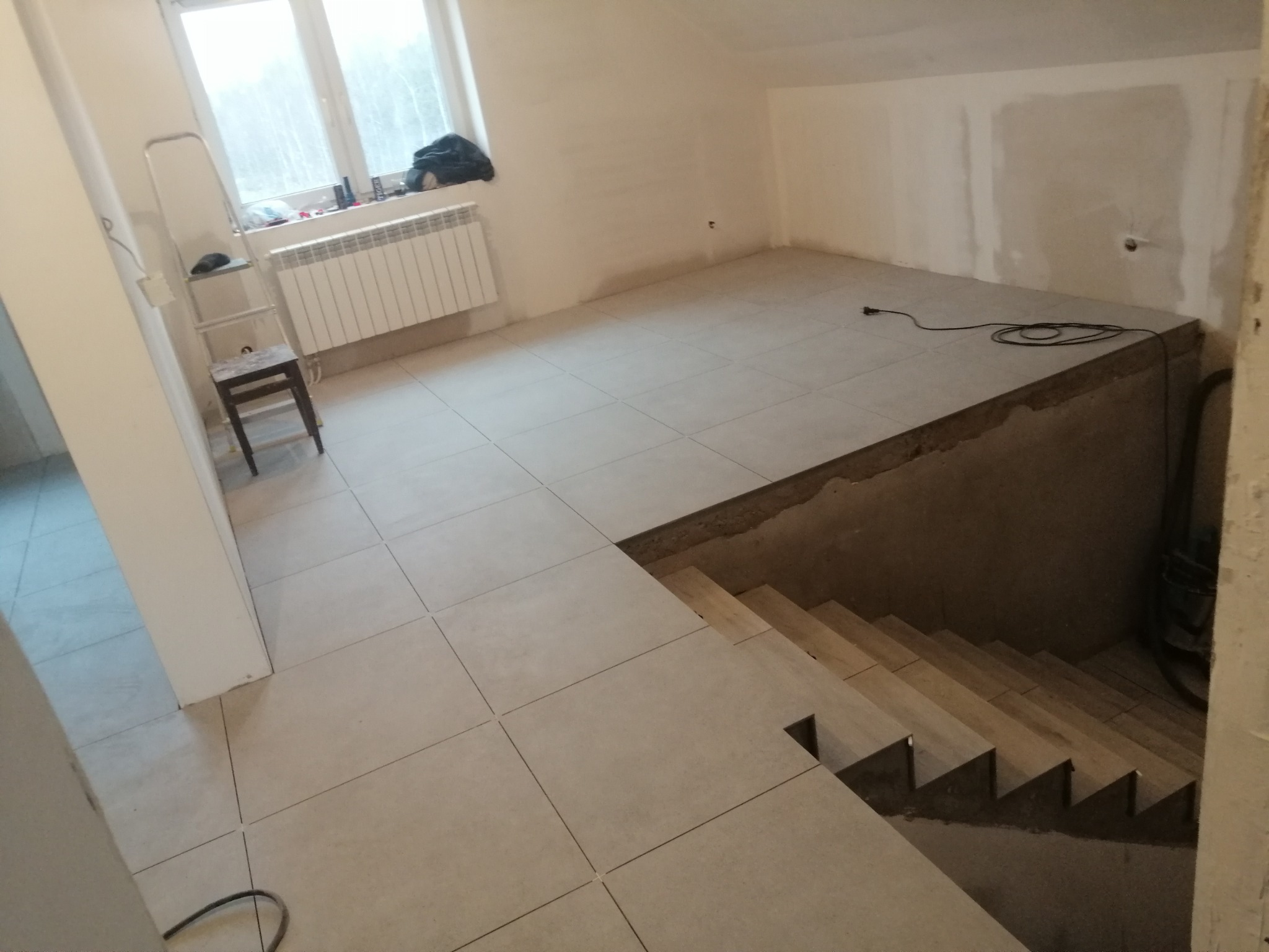 Wykończone płytki podłogowe w pomieszczeniu na poddaszu z widocznymi schodami prowadzącymi w dół, jasne ściany w trakcie przygotowania do malowania, grzejnik pod oknem.