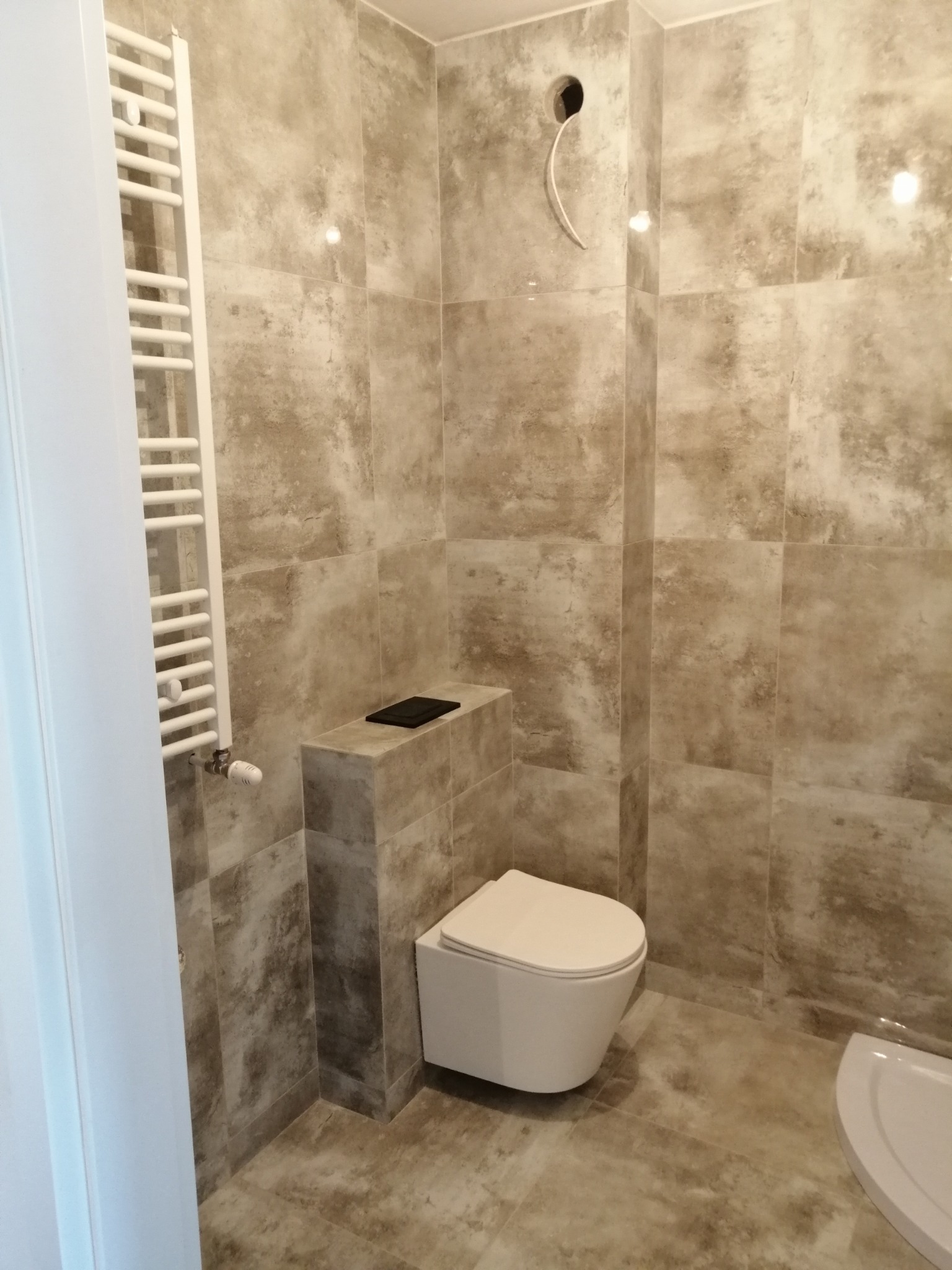 Wykończona łazienka z beżowymi płytkami imitującymi kamień, wiszącą toaletą i grzejnikiem drabinkowym po lewej stronie, z widocznym otworem na kabel w ścianie.