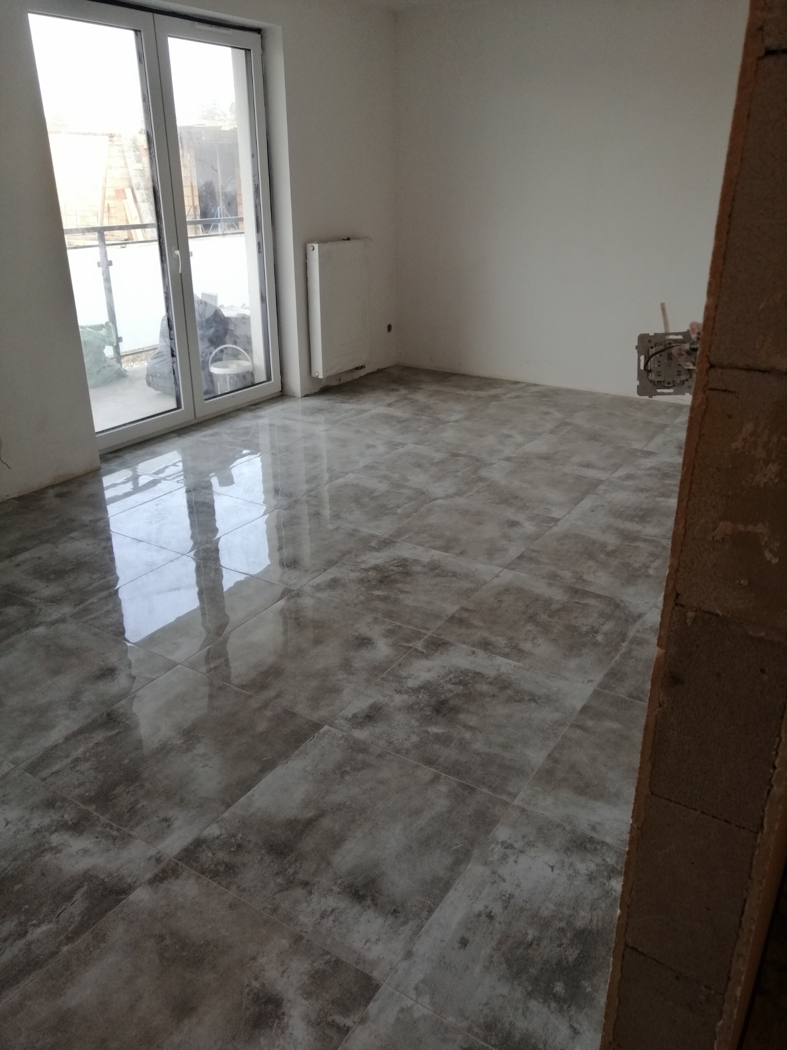 Ułożona podłoga z płytek imitujących kamień w pustym, jasnym pomieszczeniu z grzejnikiem i balkonem, widoczna instalacja elektryczna w ścianie.