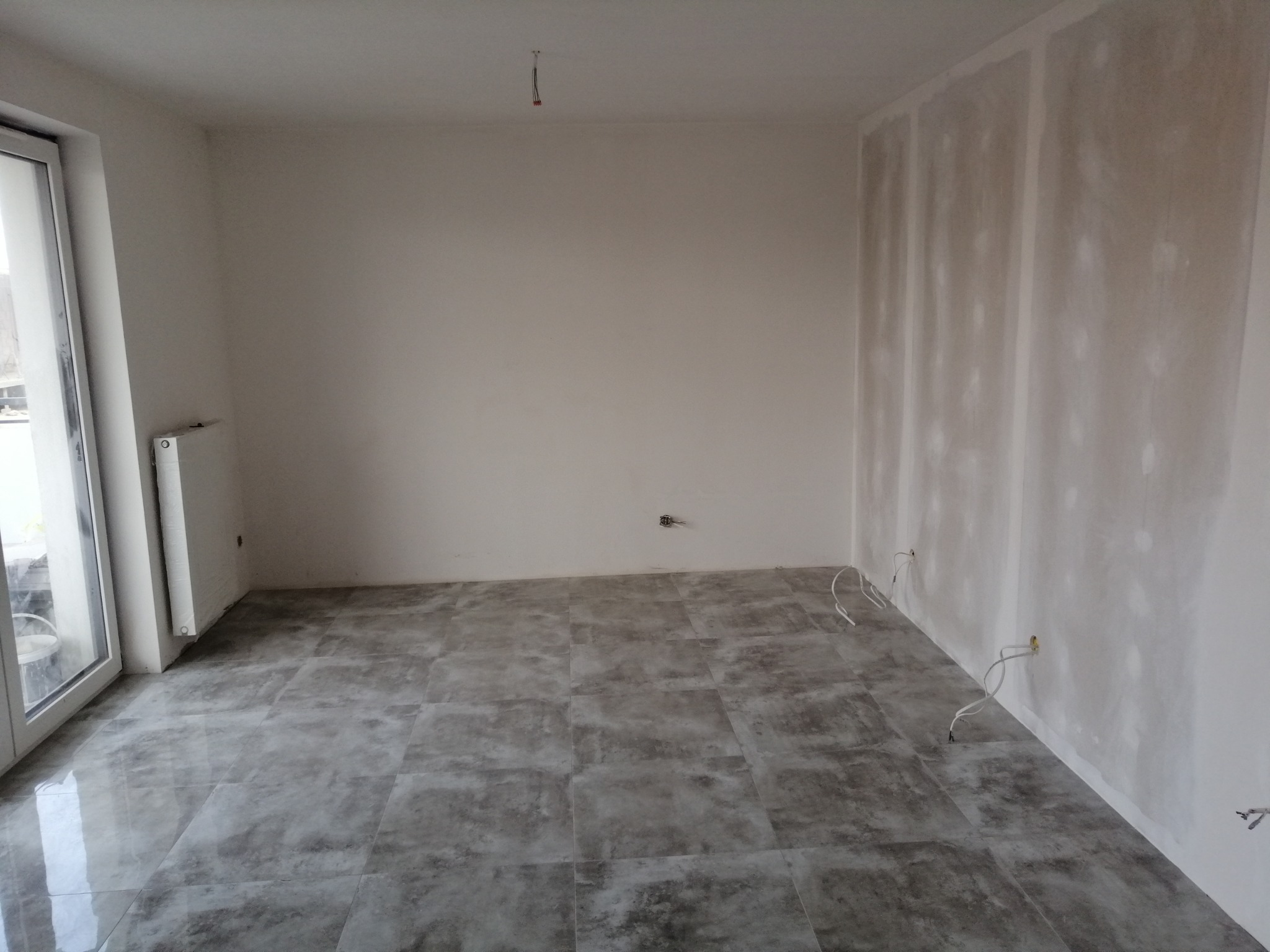 Wnętrze pokoju w trakcie prac wykończeniowych: położona szara podłoga z płytek, ściany częściowo przygotowane do malowania, widoczne wystające kable elektryczne.