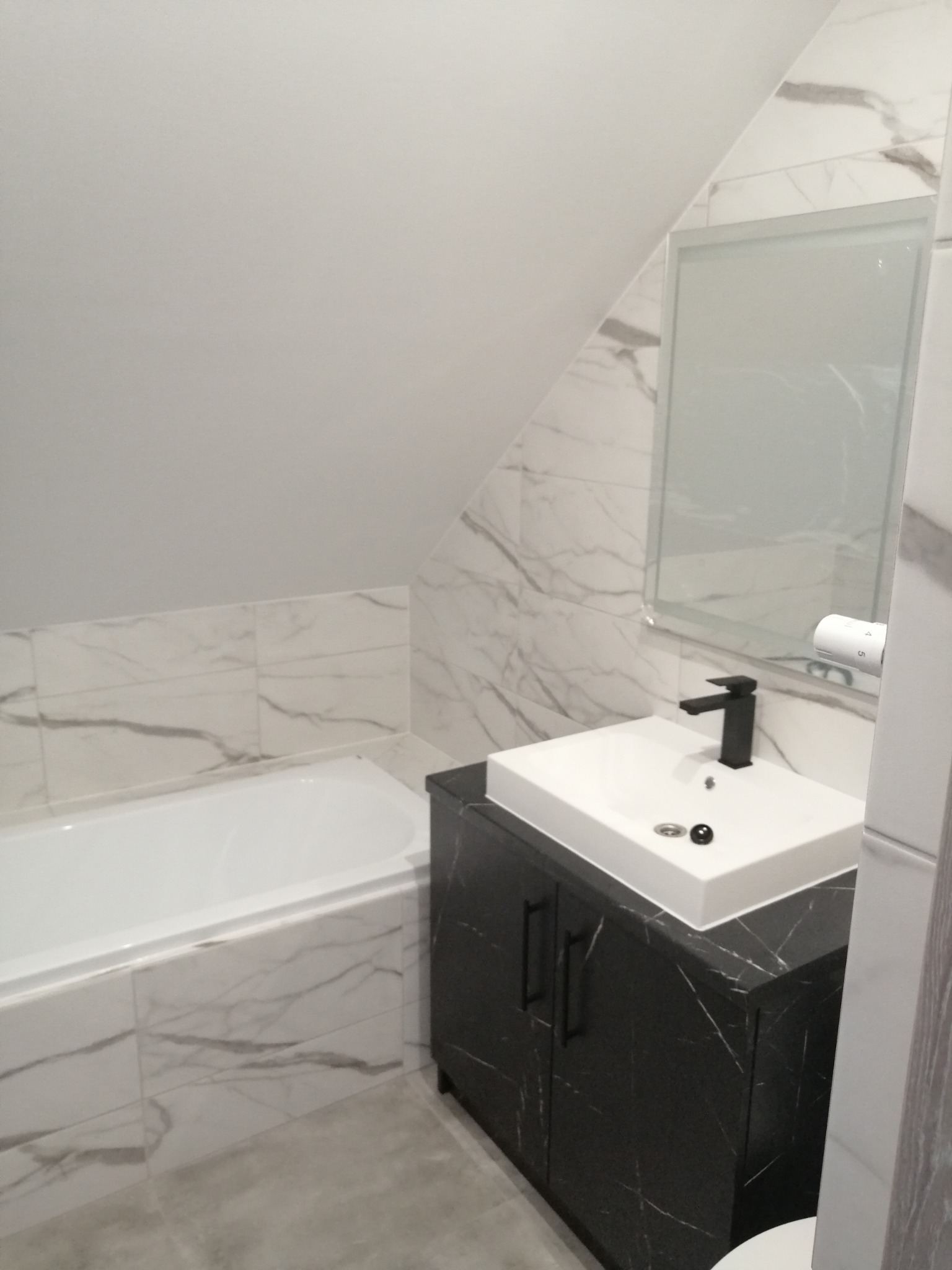 Nowoczesna łazienka na poddaszu z wanną i umywalką na szafce w kolorze czarnego marmuru, wykończona białymi płytkami imitującymi marmur.