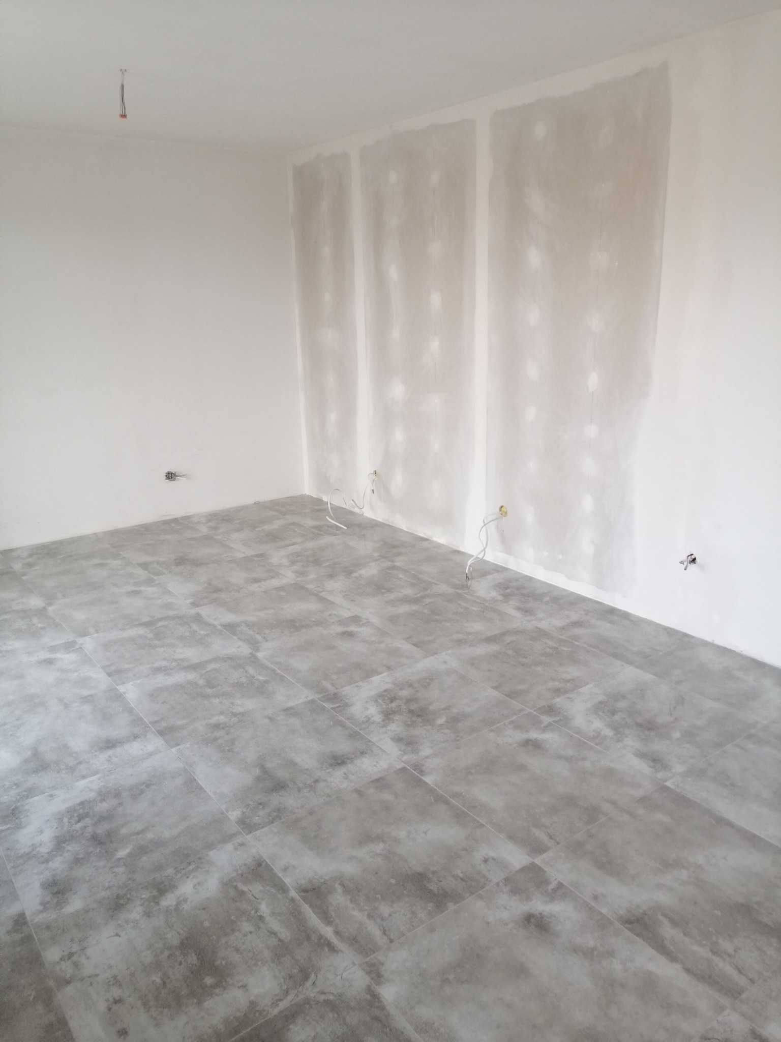 Szare płytki podłogowe imitujące beton w surowym wnętrzu z przygotowanymi ścianami do malowania i wystającymi przewodami elektrycznymi.