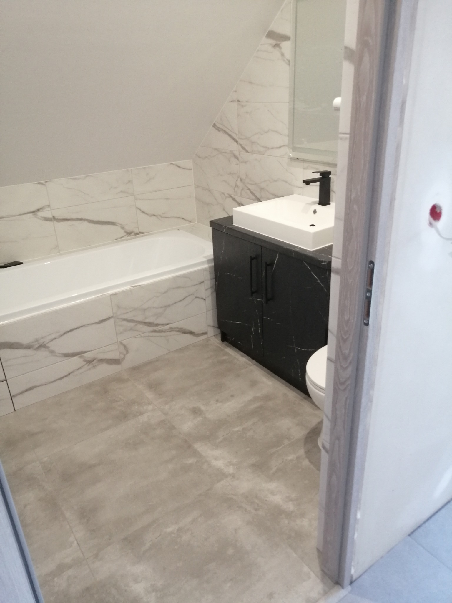 Ukończona łazienka na poddaszu z wanną obłożoną marmurowymi płytkami, umywalką nablatową na czarnej szafce i szarą podłogą z płytek, widok zza uchylonych drzwi.