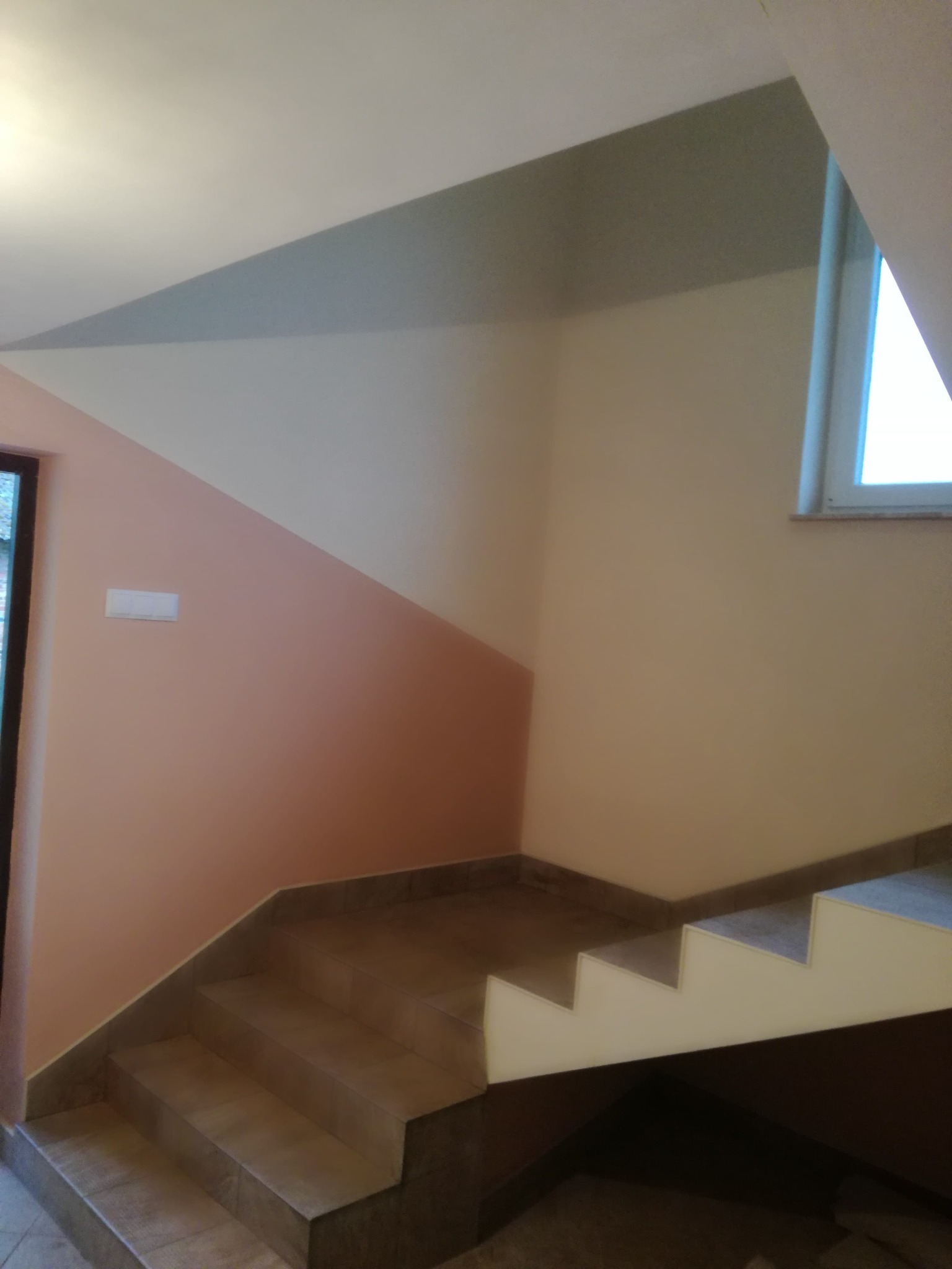 Wnętrze klatki schodowej z wykończonymi schodami i pomalowanymi ścianami w odcieniach beżu i łososia, z widocznym oknem i włącznikiem światła.