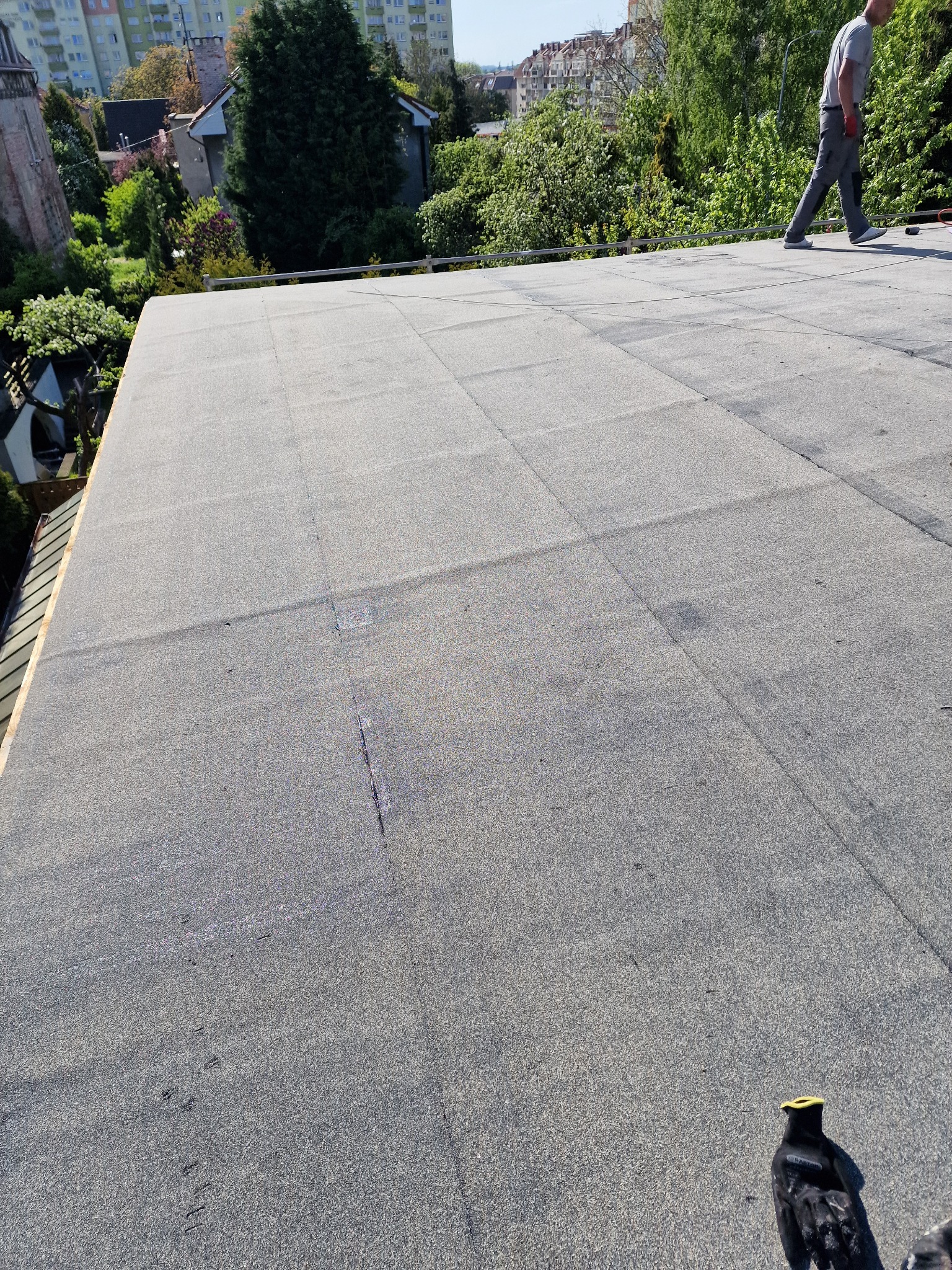 Wykończony płaski dach pokryty papą termozgrzewalną, widoczny pracownik w oddali i rękawica robocza w rogu, zieleń drzew w tle.