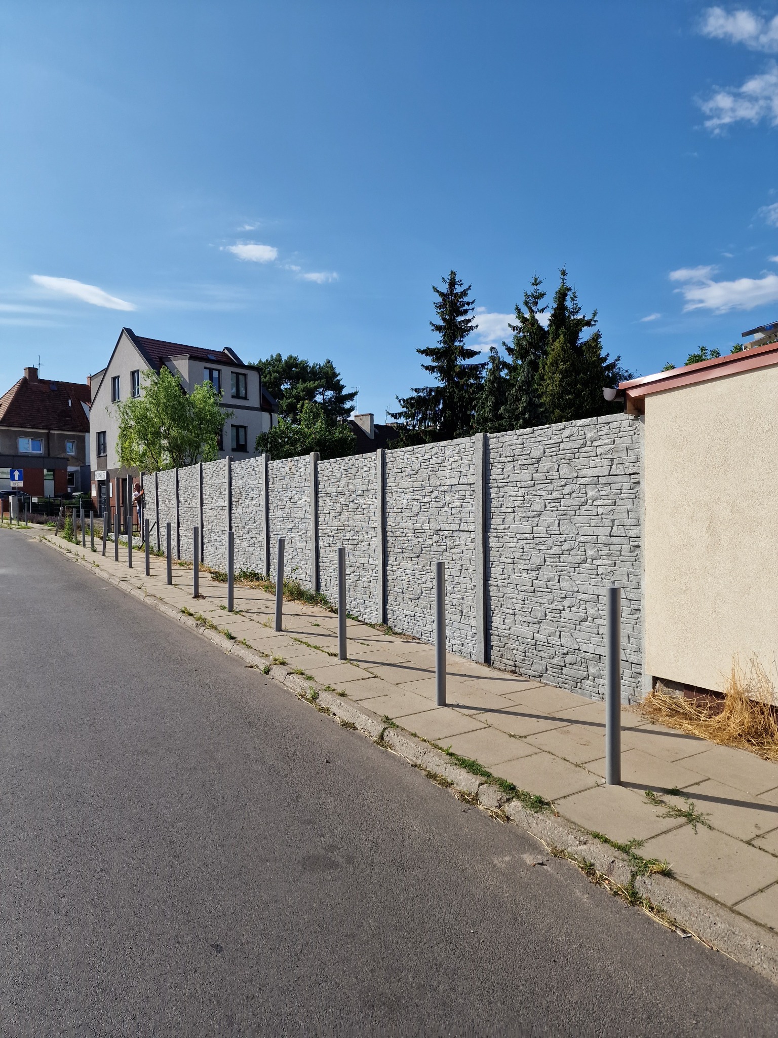 Szczecin: Długi, szary, prefabrykowany betonowy płot z fakturą kamienia, oddzielający chodnik od posesji z domami w tle, z metalowymi słupkami zabezpieczającymi.