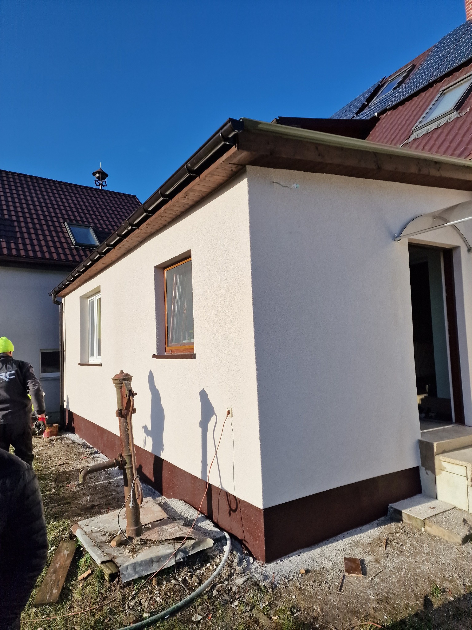 Wykończona elewacja budynku z brązowym cokołem i kremowym tynkiem, widoczne okna z drewnianymi ramami, stara studnia ręczna na pierwszym planie, pracownicy w tle.