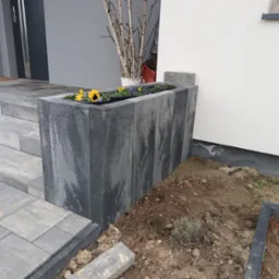 Nowoczesna donica z szarej kostki brukowej, obsadzona bratkami, umieszczona przy schodach wejściowych do domu z jasną elewacją.