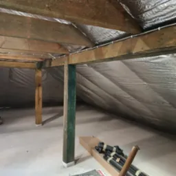 Poddasze z widoczną izolacją termiczną pokrytą folią aluminiową, drewniane belki konstrukcyjne z przymocowanymi przewodami elektrycznymi, oraz elementy instalacji w kartonie na podłodze.