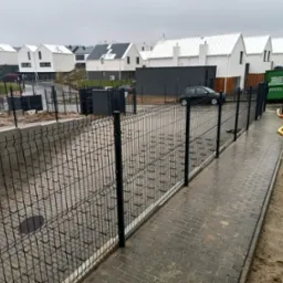 Nowo zainstalowane czarne ogrodzenie panelowe na tle szeregu nowoczesnych domów w zabudowie szeregowej, widoczne także ułożone chodniki z kostki brukowej i kontener na odpady budowlane