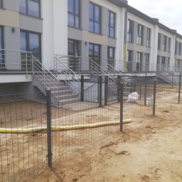 DB.Building Dominik Budzyński Usługi Wypożyczalnia - Nowoczesne segmentowe domy w zabudowie szeregowej z metalowym ogrodzeniem z siatki, widoczne żółte rury na ziemi i materiały budowlane w workach.