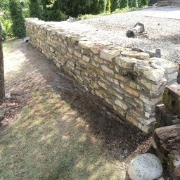 DB.Building Dominik Budzyński Usługi Wypożyczalnia - Niski mur z jasnych, nieregularnych kamieni, oddzielający trawnik od żwirowej ścieżki w ogrodzie. Widoczne drzewa i elementy drewniane.