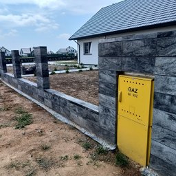 DB.Building Dominik Budzyński Usługi Wypożyczalnia - Nowoczesne ogrodzenie modułowe z szarego kamienia łupanego, z żółtą skrzynką gazową wbudowaną w przęsło. W tle dom z szarą dachówką i zadbany trawnik.