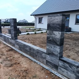 DB.Building Dominik Budzyński Usługi Wypożyczalnia - Wznoszenie ogrodzenia z szarych, betonowych bloczków. Widoczne słupki i fragment murku, w tle dom jednorodzinny i trawnik. Prace w toku, teren wokół ogrodzenia niezagospodarowany.